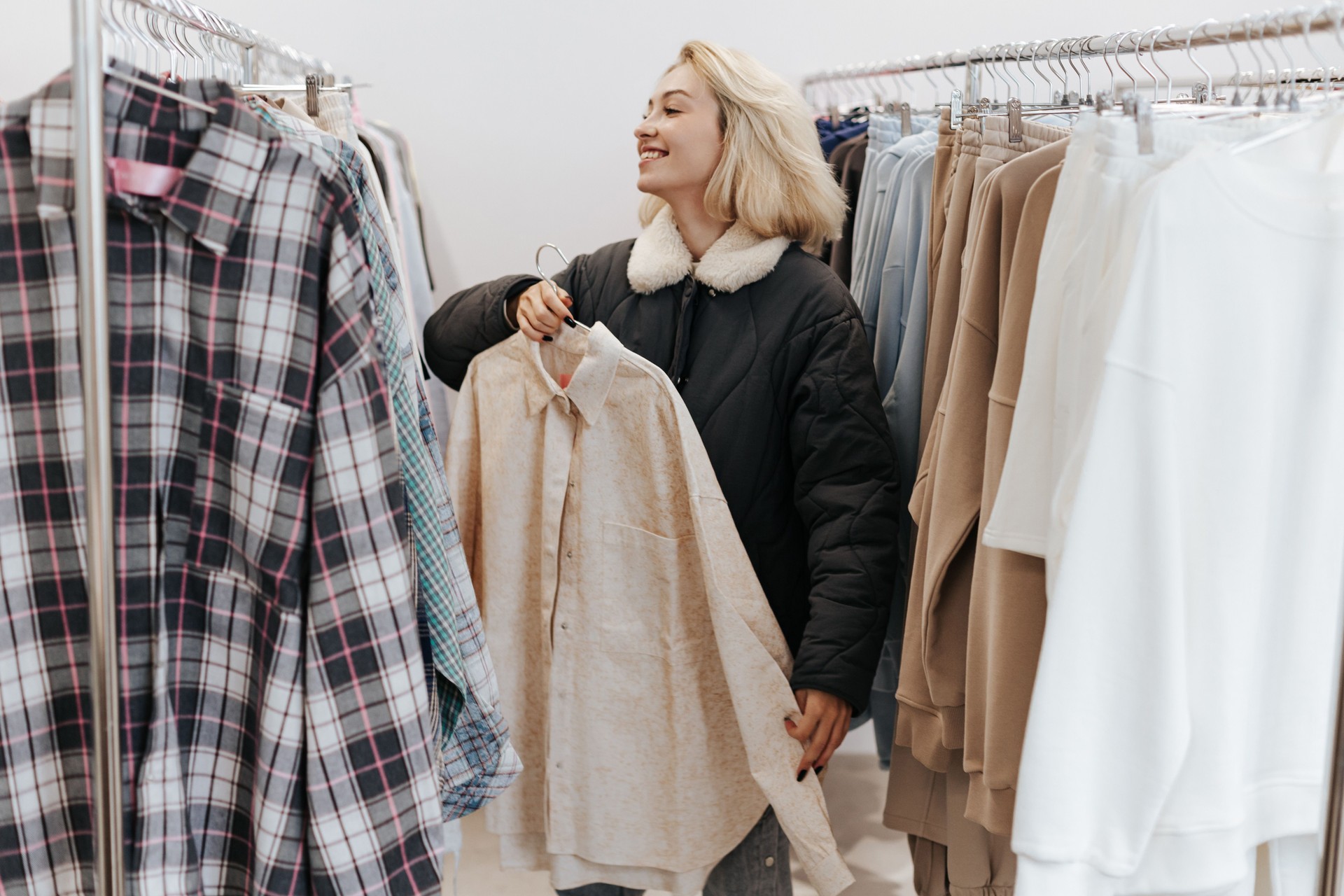 Young Smiling Blonde Woman Trying on shirt, standing between the Rails with clothes